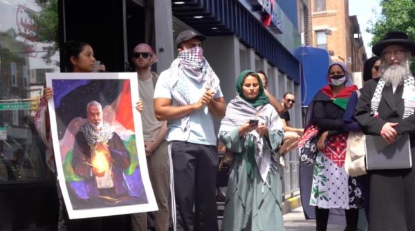 Protesters gather on the sidewalk around a photo of deceased Hamas leader Ismail Haniyeh.