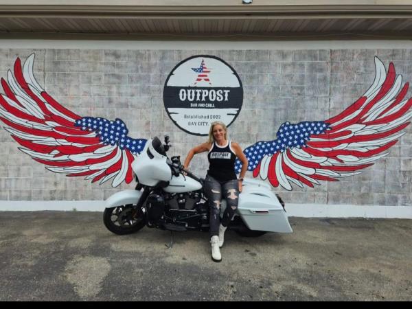 Woman in front of a motorcycle with american flag bird wings behind her on a wall.