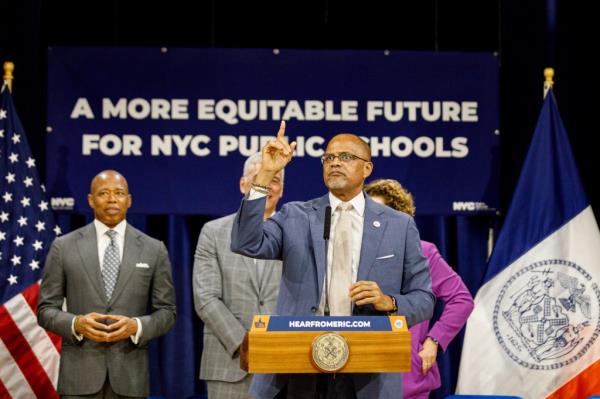 chancellor banks in a blue grey suit and yellow tie standing at a podium speaking with his right hand raised, pointing to the sky